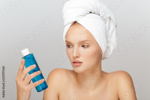 Portrait of young seriuos woman without makeup with white towel on head thoughtfully looking on blue cosmetic bottle in hand over gray background isolated