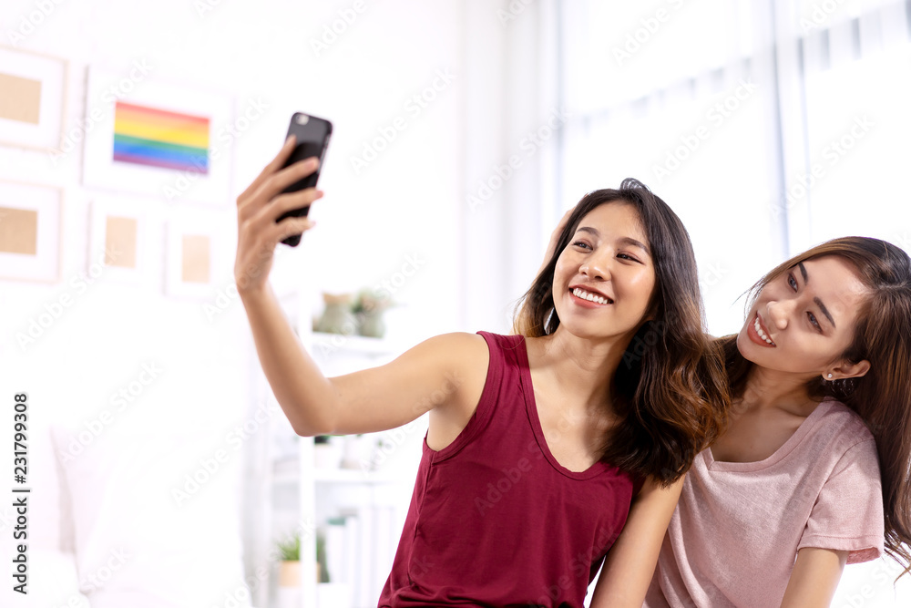 Attractive young asian lesbian student or asia girl college holding moblie phone smile and making video call or take photo to social media in white bedroom with rainbow frame background. LGBT concept. Attractive young asian lesbian student or asia girl college holding moblie phone smile and making video call or take photo to social media in white bedroom with rainbow frame background. LGBT concept.