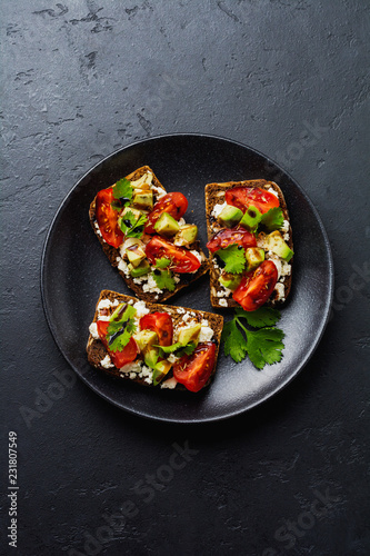 Avocados, cherry tomatoes and feta cheese sandwiches with balsamic sauce and cilantro on old olive board on dark black background. Top view.