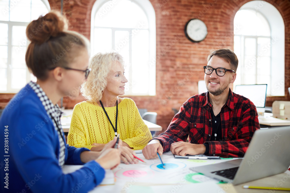 Group of creative people in casualwear sitting by table and discussing organization moments Group of creative people in casualwear sitting by table and discussing organization moments