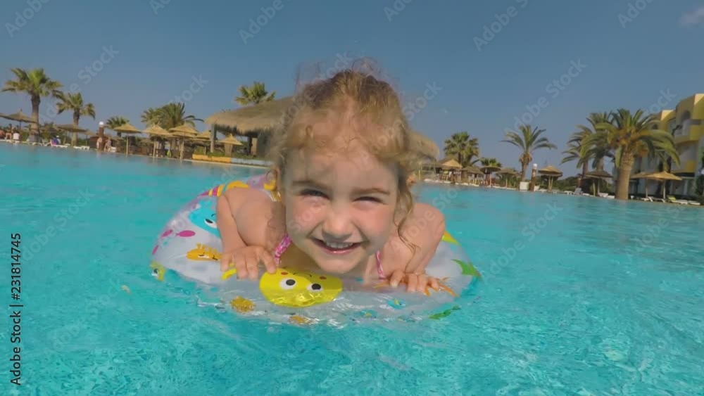 Happy little girl playing with colorful inflatable ring in outdoor swimming pool on hot summer day. Children play in tropical resort. Kids learn to swim. Family beach vacation. Happy little girl playing with colorful inflatable ring in outdoor swimming pool on hot summer day. Children play in tropical resort. Kids learn to swim. Family beach vacation.