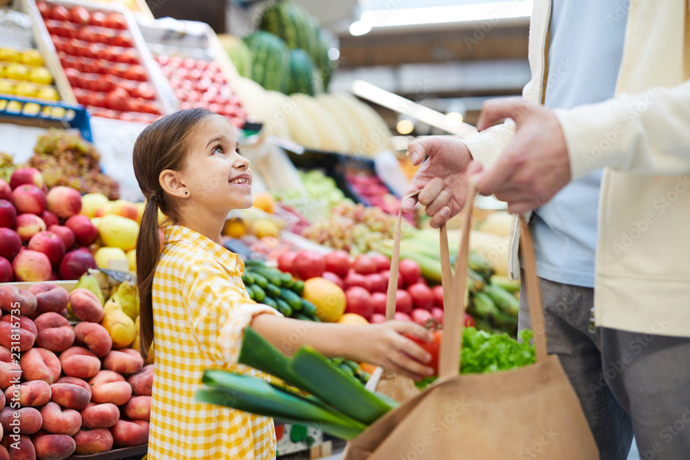 Cheerful girl in yellow dress putting vegetable into bag and looking at father while helping him with shopping at farmers market