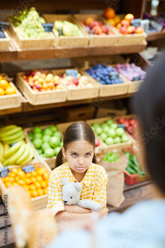 Resentful grimacing kid hugging toy bear and looking with displeasure at parent in organic food store