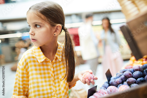 Worried girl with pony tails wearing checkered shirt standing at food shelves and holding plum while stealing it in food store photo