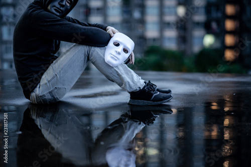 Reflection of mystery hoodie man with black mask holding white mask sitting in the rain on rooftop of abandoned building. Bipolar disorder or Major depressive disorder. Depression concept