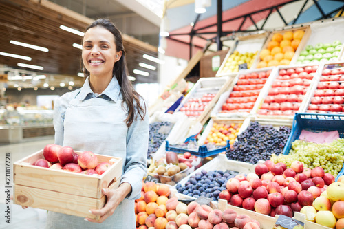 Cheerful excited young female grocery retailer in apron standing against food stall and holding box of apples