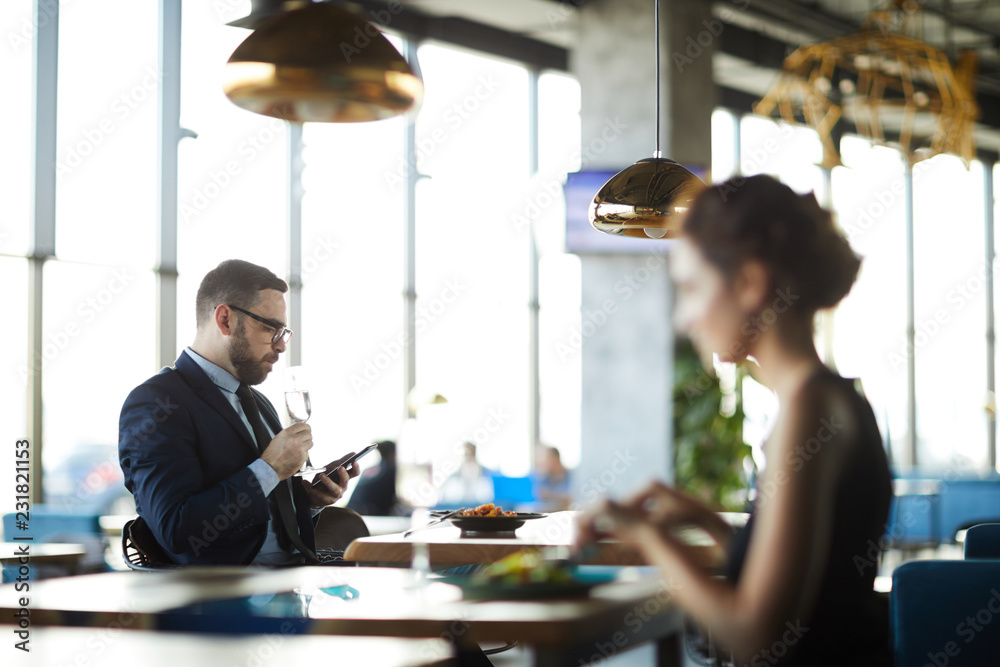 Young contemporary businessman with flute of champagne texting in smartphone while sitting by table in restaurant Young contemporary businessman with flute of champagne texting in smartphone while sitting by table in restaurant