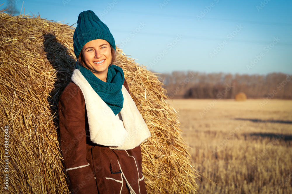 Young attractive lady in blue knitting hat and brown coat stay under haystack. Smiling beautiful hipster happy woman on field, wearing stylish clothes. Autumn trend, urban stydent style. Young attractive lady in blue knitting hat and brown coat stay under haystack. Smiling beautiful hipster happy woman on field, wearing stylish clothes. Autumn trend, urban stydent style.