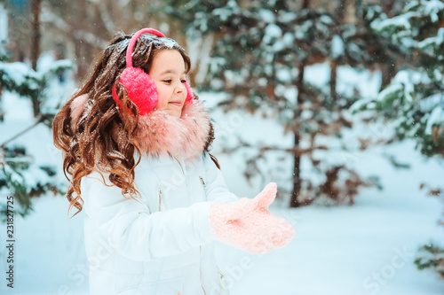 Winter portrait of happy kid girl in pink earmuffs walking outdoor in snowy winter forest. Happy childhood and active winter holidays concept