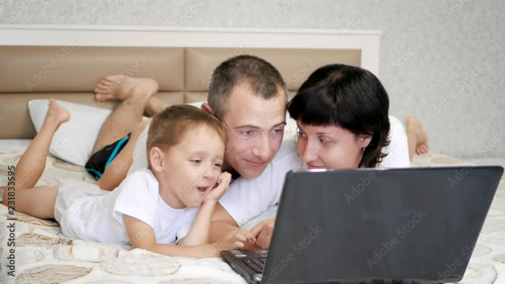 Happy family: dad, mom and baby are lying on the bed and using a laptop, smiling. Happy family: dad, mom and baby are lying on the bed and using a laptop, smiling.