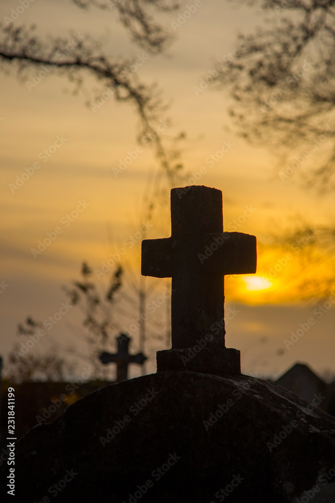 a cross on the background of the sunset and branches of the trees, a mysterious atmosphere. A weathered stone Christian cross silhouetted against an orange sunset.