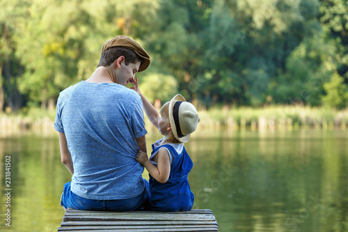 Father and a tiny daughters in dungarees are sitting on a river bridge and looking at each other. They talk, laugh and have fun.