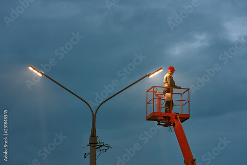 Municipal worker with helmet and safety protective equipment installs new diode lights. Worker in lift bucket repair light pole. Modernization of street lamps. Technician on aerial device. Night duty