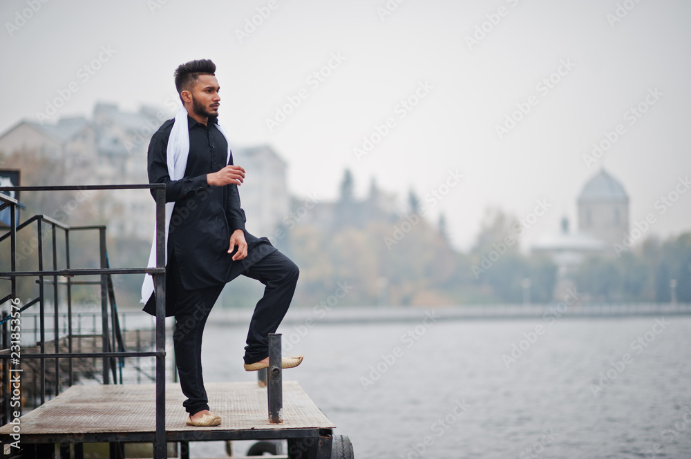 Indian stylish man in black traditional clothes with white scarf posed outdoor against foggy pier of the sea. Indian stylish man in black traditional clothes with white scarf posed outdoor against foggy pier of the sea.