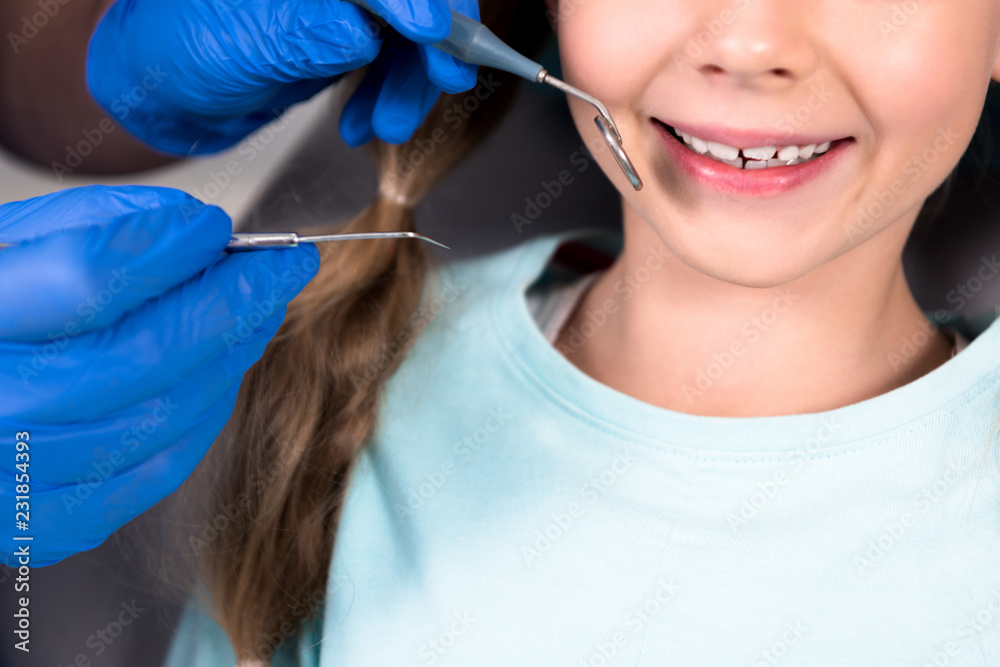 cropped shot of dentist with tools examining teeth of smiling little child cropped shot of dentist with tools examining teeth of smiling little child