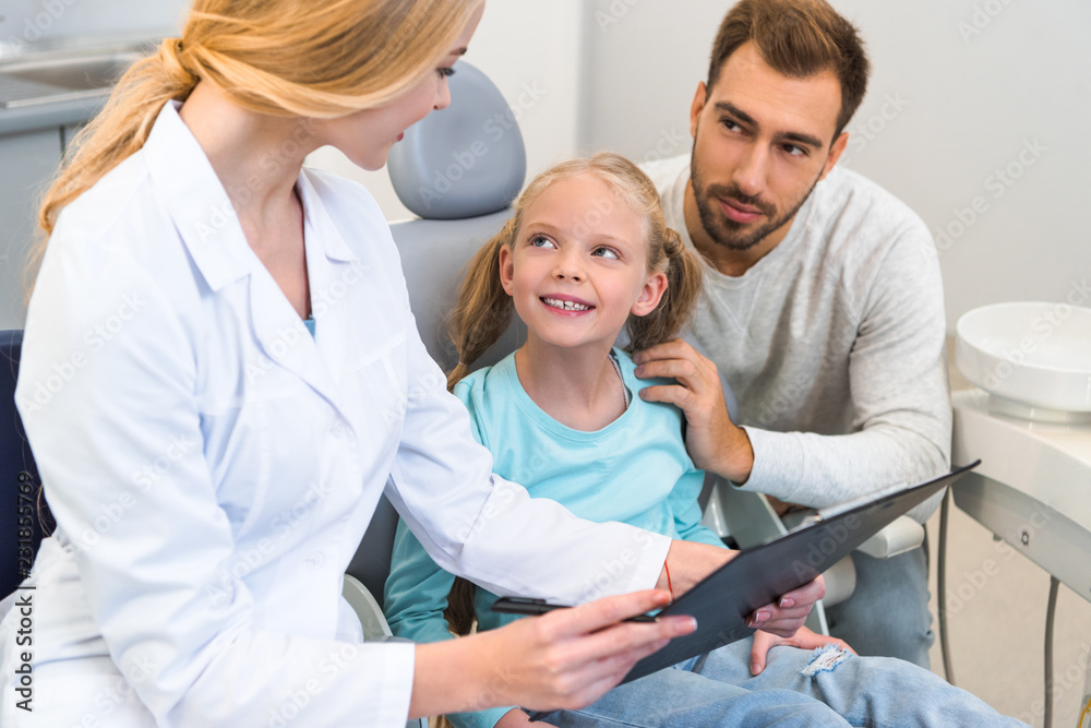 Obraz premium young female dentist showing clipboard with diagnosis to father and little daughter
