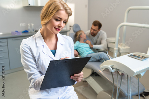 female dentist writing in clipboard while little child and her father sitting on background