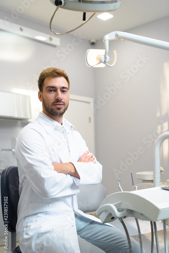 handsome young dentist with crossed arms looking at camera in office