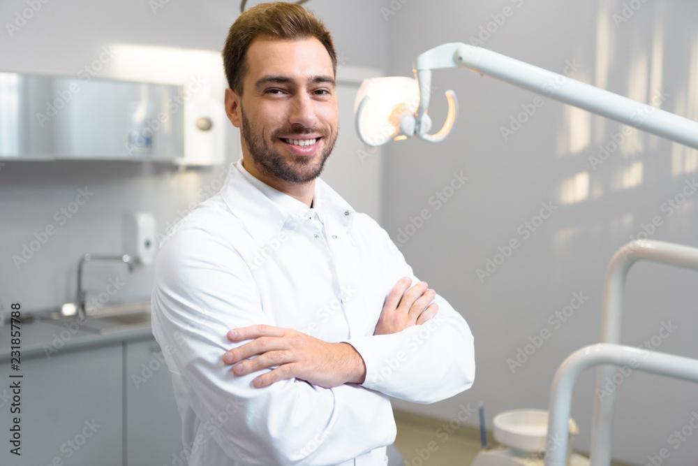 handsome young dentist with crossed arms looking at camera in office