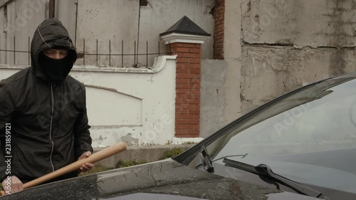 Aggressive vandal in mask and hood going to crash car with baseball bat on street
