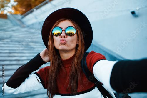 Kiss me! Beautiful young woman making selfie on camera.Gorgeous beauty. Self portrait of attractive young woman looking at camera and sending kiss while standing outdoors,wearing hat and sunglasses.