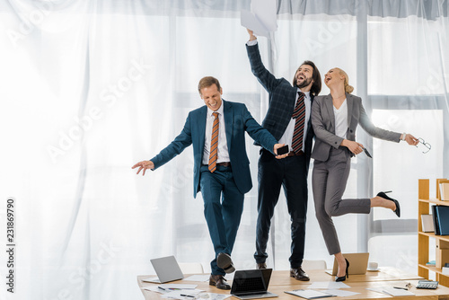 joyful insurance workers dancing on table and throwing papers at meeting in office