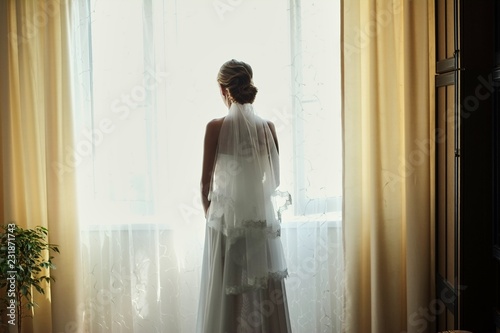 Beautiful bride in white wedding dress standing in her bedroom and looking in window