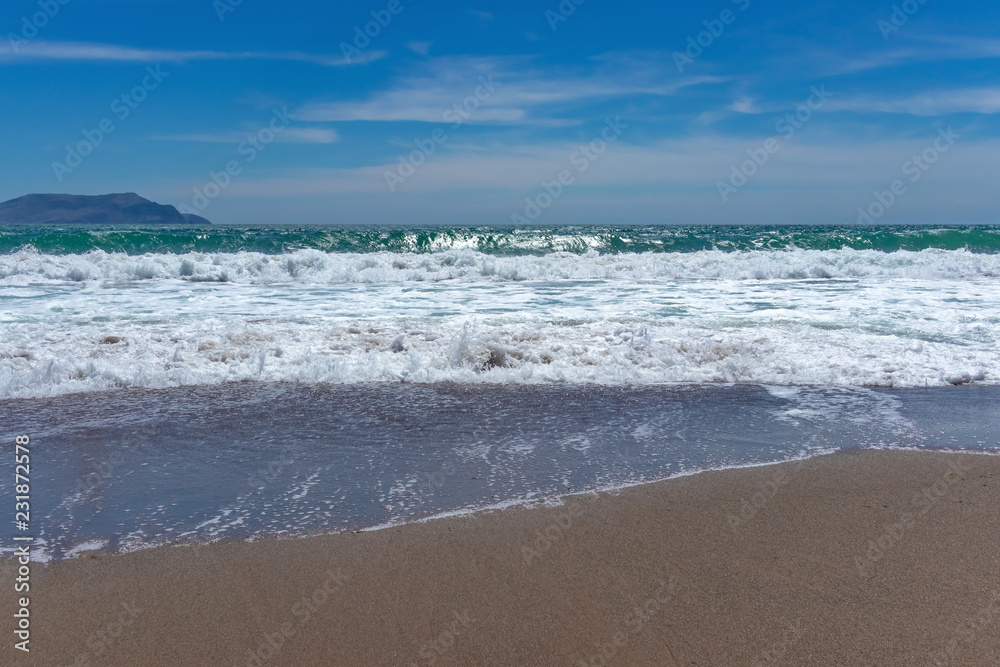 Beautiful turquoise waves with foam on a sandy beach, against the blue sky with clouds and a mountain on the horizon