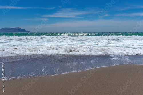 Beautiful turquoise waves with foam on a sandy beach, against the blue sky with clouds and a mountain on the horizon