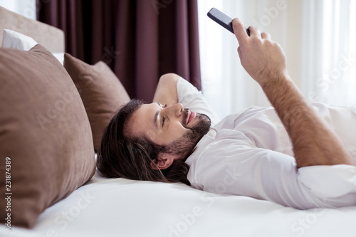 Digital device. Joyful happy man using his modern smartphone while lying on the bed