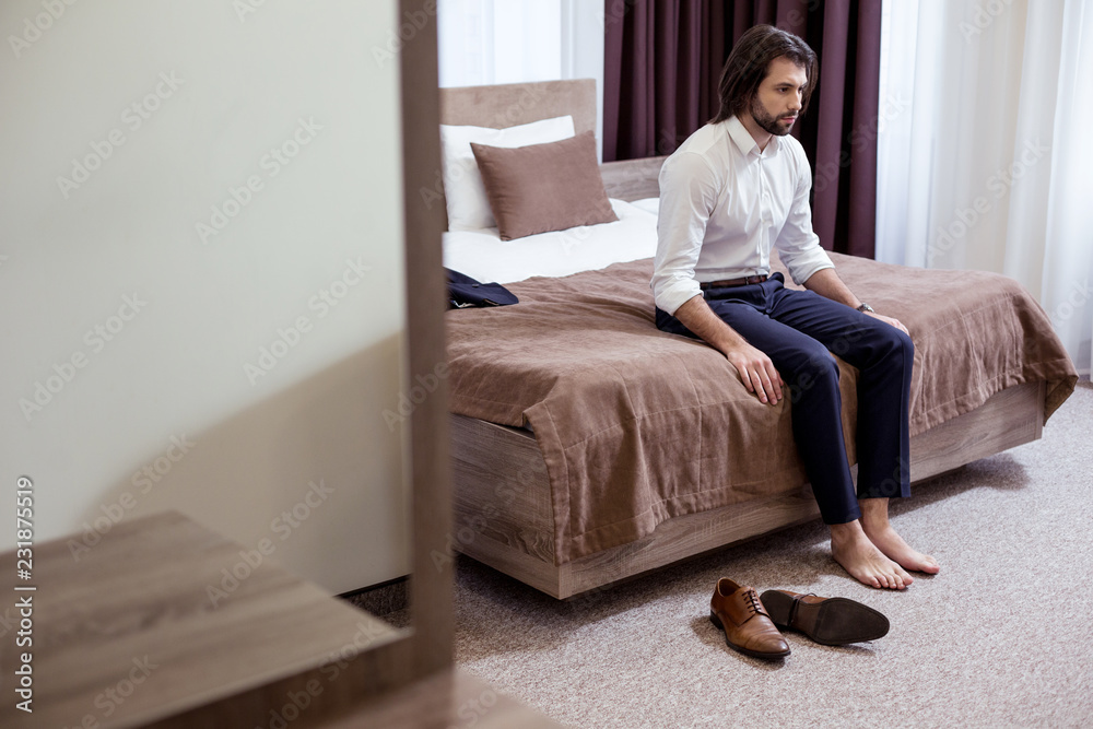 In the hotel room. Sad tired man sitting on the bed in the hotel while being very tired In the hotel room. Sad tired man sitting on the bed in the hotel while being very tired