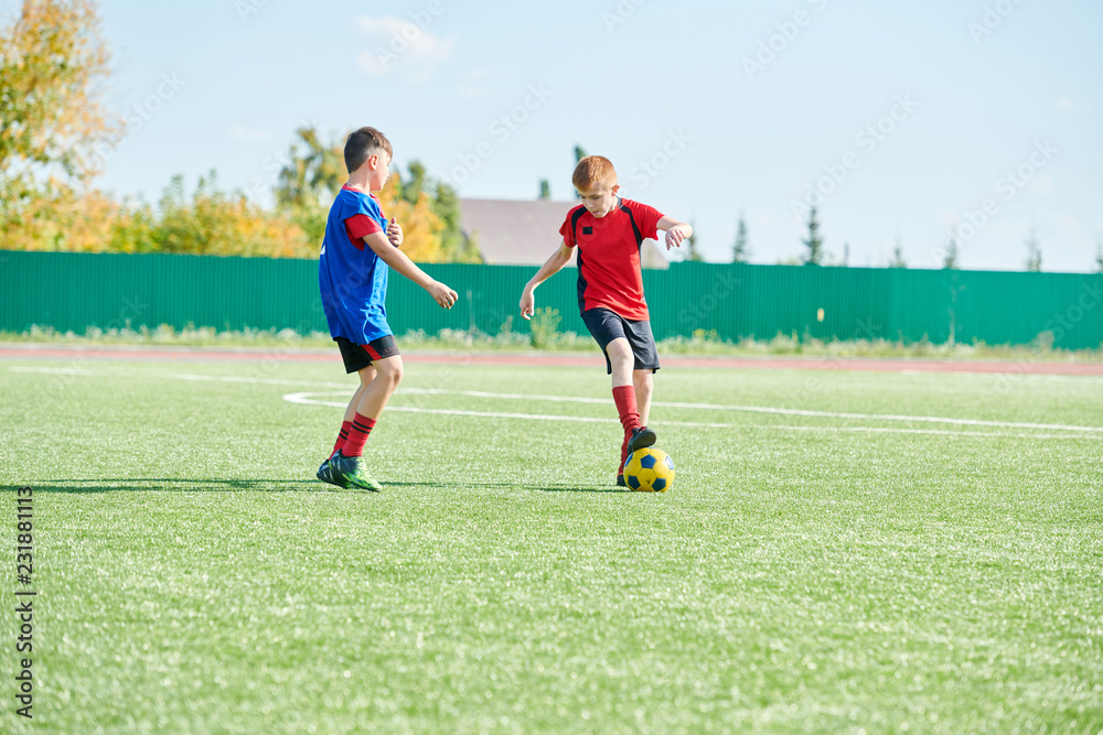 Obraz premium Full length portrait of two boys playing football in outdoor stadium lit by sunlight, copy space