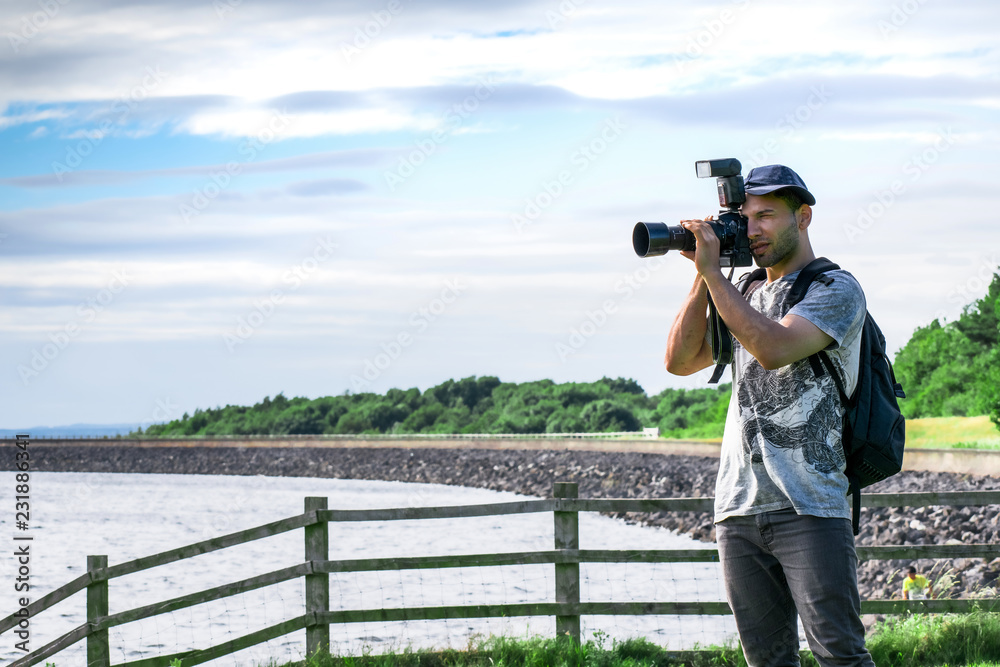 Closeup of Young handsome Professional photographer in white shirt with black backpack, taking landscape photos on the beach close to a wooden fence in a green nature with blue cloudy sky
