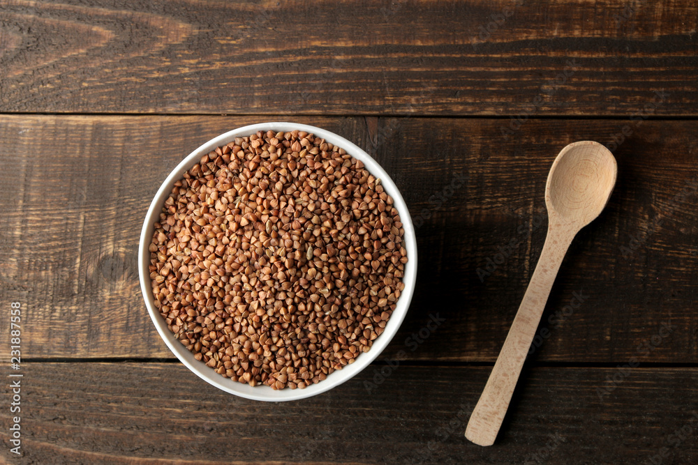 Dry buckwheat groats in a white bowl with a spoon on a wooden brown table. cereals. healthy food. porridge. top view Dry buckwheat groats in a white bowl with a spoon on a wooden brown table. cereals. healthy food. porridge. top view
