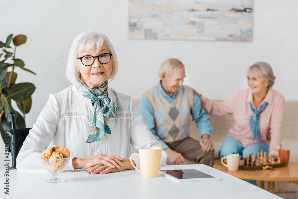 senior woman in glasses drinking tea with cookies while senior couple playing chess senior woman in glasses drinking tea with cookies while senior couple playing chess