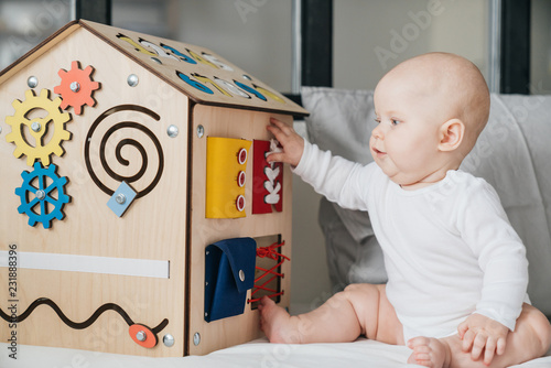 Busy Board. A special toy house model with many functions for the development of fine motor skills. A child plays with a wooden house toy sitting on a bed by the window.