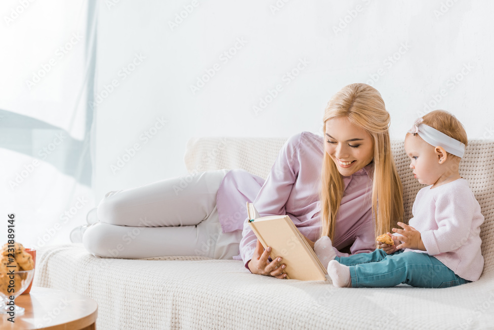 young smiling mother reading book to small daughter while toddler holding cookie young smiling mother reading book to small daughter while toddler holding cookie