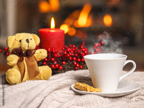 Cup of hot drink with steam and teddy bear, cookie berries red candle in Christmas decoration on knitted plaid in front of fireplace. Christmas New Year concept. Cozy relaxed atmosphere home interior.