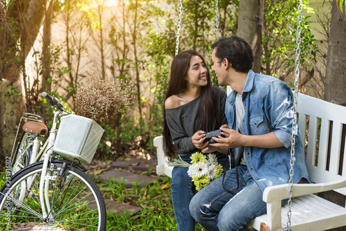 Asian couple having eye contact as romantic moment at bench in natural park with bicycle. People and lifestyles concept for Valentines day and wedding ceremony. Love at first sight theme.