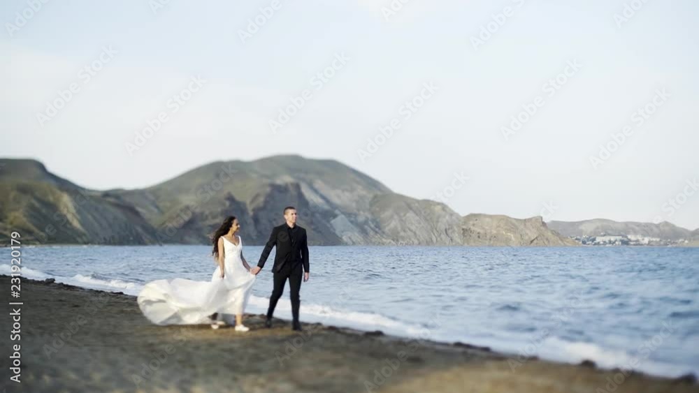 Newlywed man and woman walking on sandy beach at background blue calm sea and beautiful landscape Newlywed man and woman walking on sandy beach at background blue calm sea and beautiful landscape