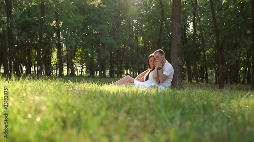 Young teen lovers are sitting on the grass leaning against a tree in the Park in Sunny warm weather. Wide shot. Slow motion. photo