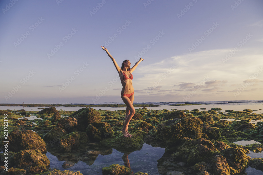 Young woman in a red bikini with raised hands at the beach, standing on the moss covered rocks, full-length portrait with natural sunlight, wellbeing holiday concept
