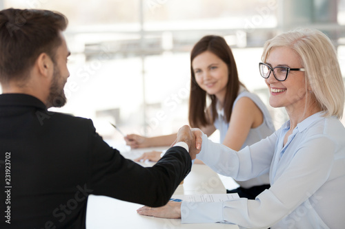 Diverse company workers handshake at briefing in office, smiling mature woman shake hand of millennial male colleague, get acquainted during business meeting, coworkers greeting introducing