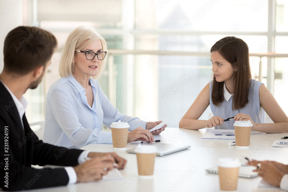 Diverse employees discuss project statistics at company business meeting, millennial employees listen to mature businesswoman talk, middle aged female boss negotiate with young coworkers at briefing