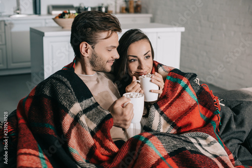 happy young couple with mugs of cocoa with marshmallow relaxing on couch and covering with plaid