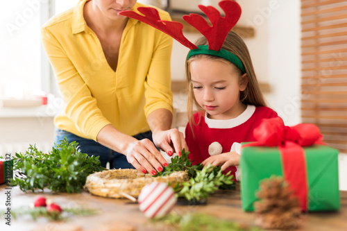 Cute preschooler girl wearing reindeer antlers and her mother making christmas wreath in living room. Christmas decoration family fun concept.