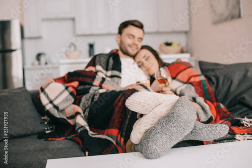 happy young couple with glasses of white wine relaxing on couch under plaid