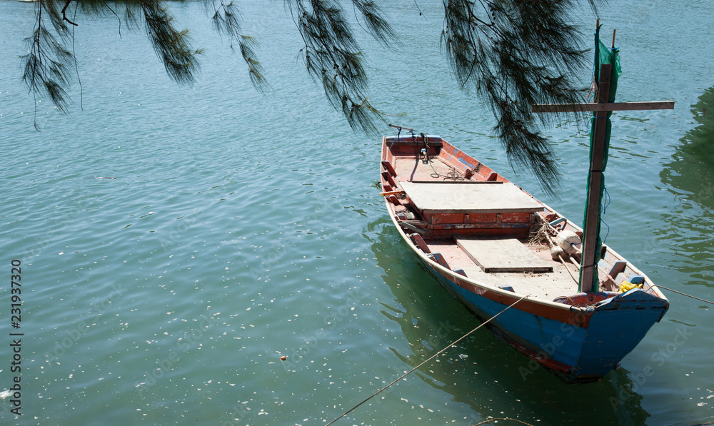 fisherman fishing boat anchored at pier under pine tree with natural blue green seawater background. marine vessel, occupational tool and equipment, transportation, agriculture or fishery concept.