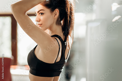 Fototapeta Naklejka Na Ścianę i Meble -  Beautiful slim girl dressed in black sportswear is standing in the gym and tying her hair