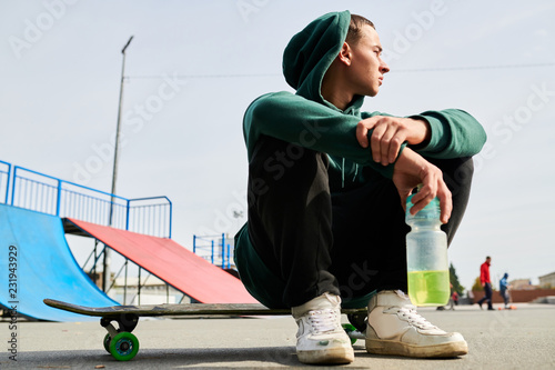 Full length portrait of unrecognizable teenager sitting on skateboard while chilling at extreme park outdoors, copy space